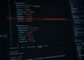 A close-up angled monitor displaying JavaScript dependency files, npm audit logs, and developer tooling interfaces with subtle red vulnerability warnings highlighted across the screen, creating a realistic software supply chain security atmosphere in a dark minimal workspace.