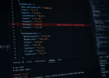 A close-up angled monitor displaying JavaScript dependency files, npm audit logs, and developer tooling interfaces with subtle red vulnerability warnings highlighted across the screen, creating a realistic software supply chain security atmosphere in a dark minimal workspace.